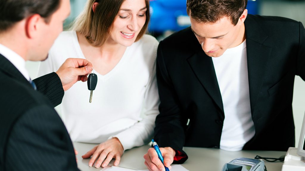 A Smiling customer couple signing financing paperwork for a new Buick SUV purchase at a dealership as the sales rep presents them with the keys